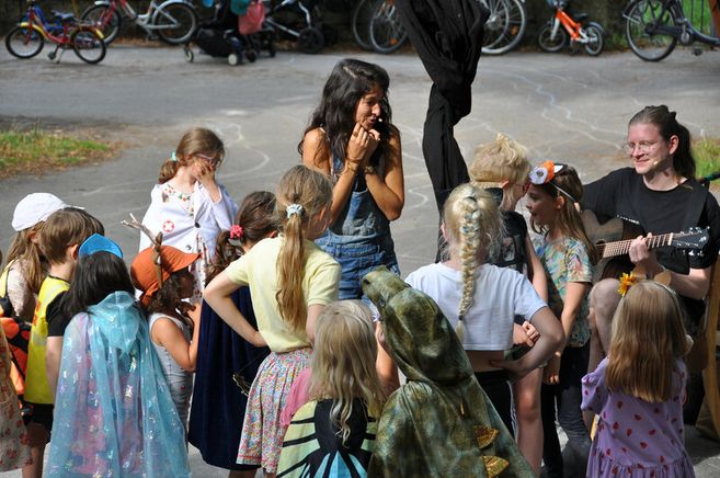 Kinder in Kostümen stehen im Kreis um eine Frau und einen Gitarrenspieler im Freien.