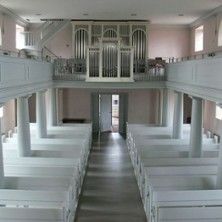 Innenansicht einer Kirche mit weißen Holzbänken und einer Orgel auf der Empore.