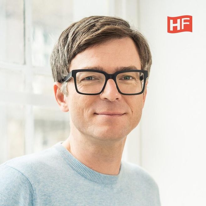 Porträt eines Mannes mit Brille und hellblauem Pullover vor einem Fenster.