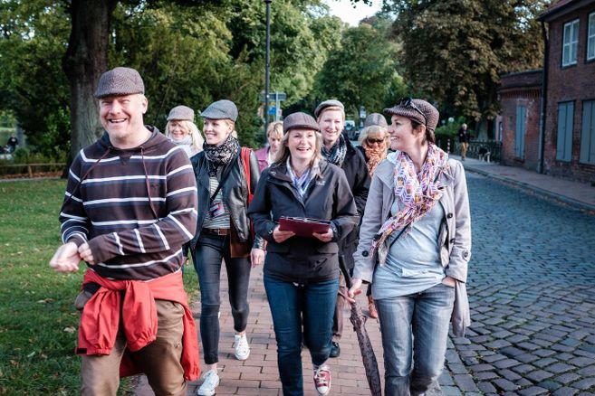 Eine Gruppe von Menschen mit Hüten geht lächelnd auf einem gepflasterten Weg in einer grünen Umgebung.