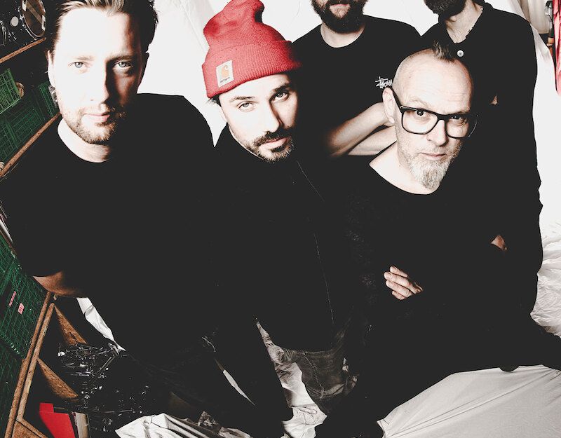 Fünf Männer in schwarzer Kleidung posieren in einem Raum mit weißen Wänden.