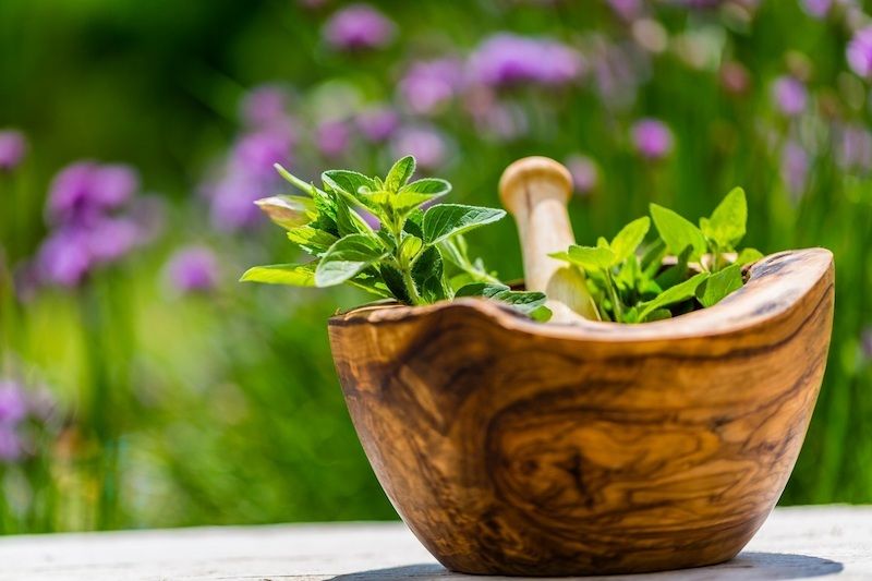 Ein hölzerner Mörser mit frischen Kräutern im Freien, umgeben von unscharfen lila Blumen im Hintergrund.