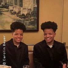 Zwei lächelnde Personen in dunkler Kleidung sitzen in einem Restaurant vor einem Gemälde.