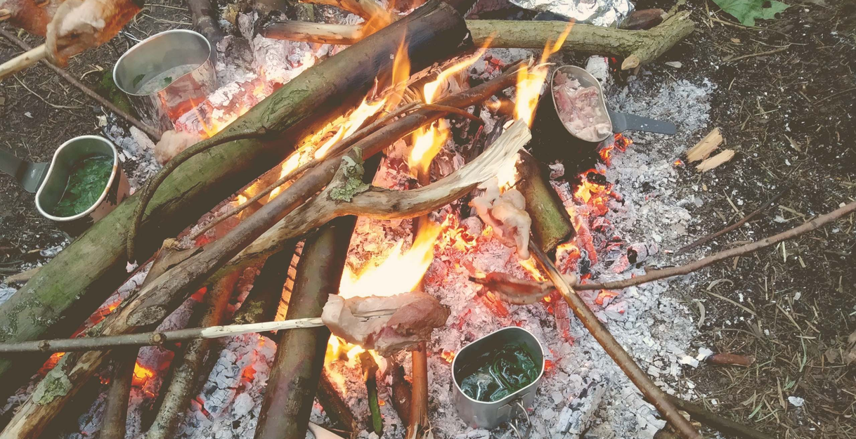 Lagerfeuer mit Kochutensilien und Fleisch am Spieß.