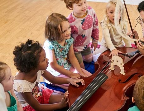 Kinder sitzen um ein Cello, während eine erwachsene Person ihnen etwas zeigt.