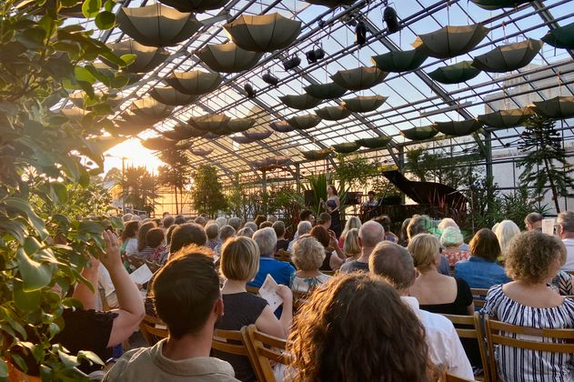 Blick in das Gewächshaus 1 des Loki-Schmidt-Gartens bei einem Konzert. Im Vordergrund viele Sitzreihen in der RÜckenansicht, im Mittelgrund ein aufgeklappter Konzertflügel, am oberen Bildrand die Gläserne Decke, an der zur Verschattung umgedrehte Regenschirme hängen