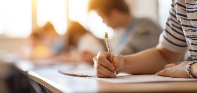 Nahaufnahme einer Person, die mit einem Stift eine Prüfung oder Klassenarbeit schreibt, im unscharfen Hintergrund weitere sitzende Teilnehmende in einem Unterrichtsraum