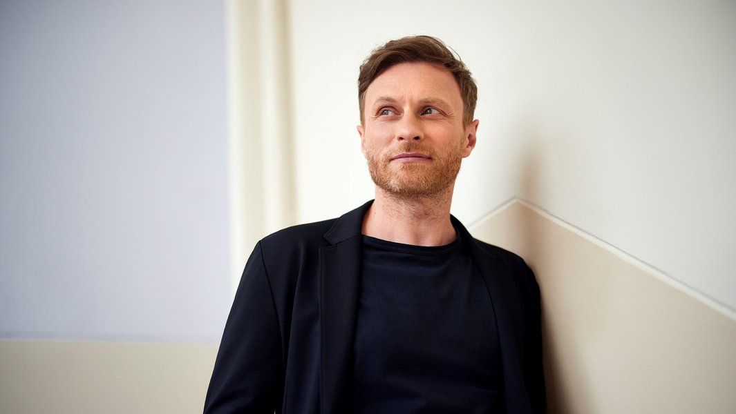 Ein Mann in schwarzer Kleidung lehnt an einer Wand und schaut nach rechts.