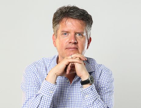 Ein Mann mit grauem Haar sitzt in einem karierten Hemd. Er stützt sein Kinn auf ineinandergelegte Hände.