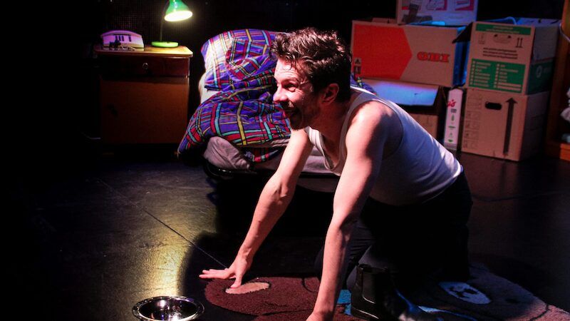 Ein Mann kniet auf einem Teppich in einem Raum mit einem Bett und Kartons im Hintergrund.