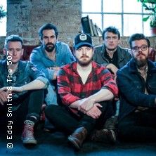 Eine Band sitzt in einem Raum mit Backsteinwand und Fenster im Hintergrund.