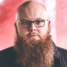 Ein Mann mit Brille und langem Bart vor einem verschwommenen roten Hintergrund.