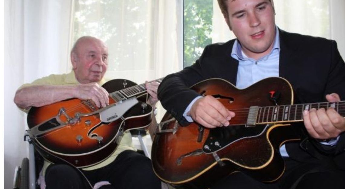 Zwei Männer spielen Gitarre, einer älter, einer jünger, vor einem Fenster mit weißen Vorhängen.