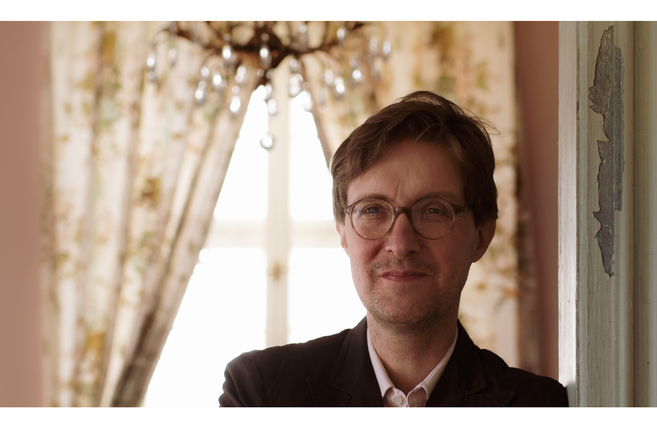 Ein Mann mit Brille steht vor einem Fenster mit Vorhängen und einem Kronleuchter im Hintergrund.
