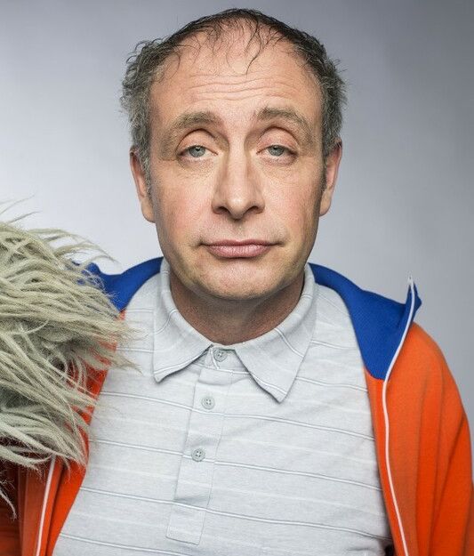 Ein Mann mit grauem Hemd und orange-blauer Jacke schaut direkt in die Kamera. Neben ihm ist ein graues, zotteliges Objekt zu sehen.