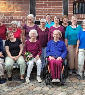 13 Frauen des Chores "FrauenTöne" und Chorleiterin stehen in 2 Reihen vor einem Fahwerkhaus. Sie tragen helle Hosen und Oberteile in Blau- und Rottönen.