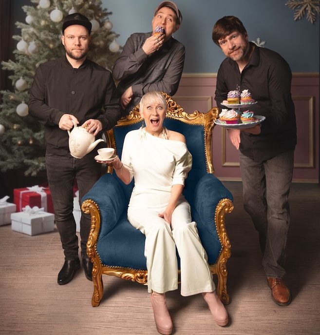 Vier Personen posieren vor einem Weihnachtsbaum. Eine Frau sitzt auf einem blauen Sessel, während drei Männer um sie herum stehen und Tee und Cupcakes halten.