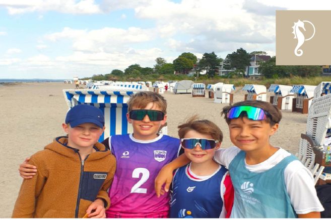 Vier Kinder stehen am Strand vor Strandkörben und lächeln in die Kamera. Sie tragen sportliche Kleidung und Sonnenbrillen.