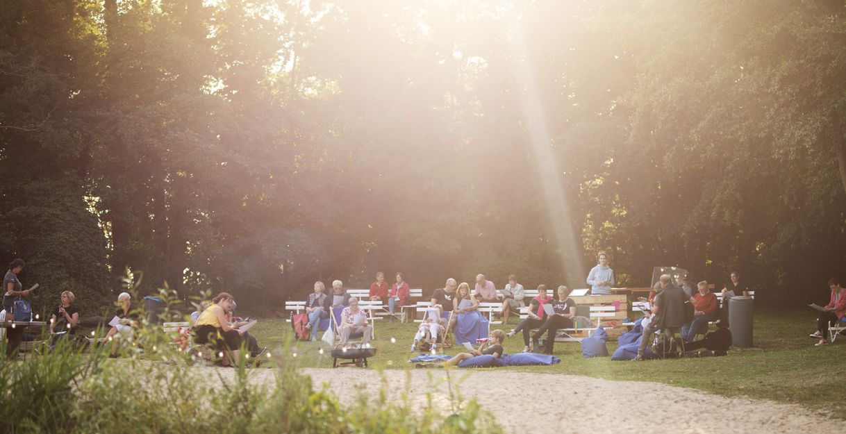 Menschen sitzen im Freien auf Bänken und Stühlen, umgeben von Bäumen, während Sonnenlicht durch die Blätter scheint.