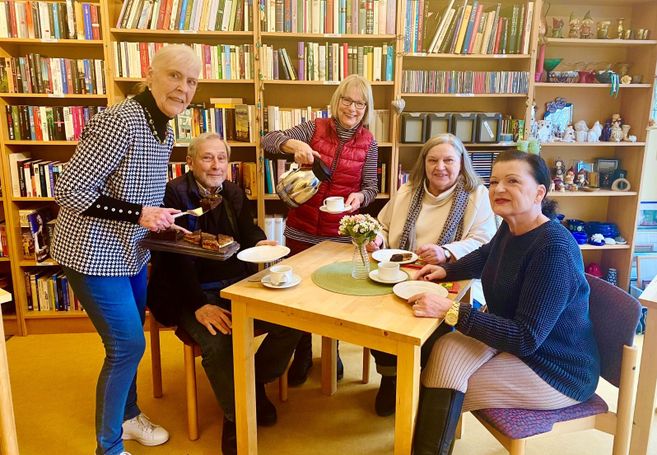 Eine Gruppe älterer Menschen sitzt an einem Tisch in einem Café mit Bücherregalen im Hintergrund. Eine Frau serviert Kuchen, eine andere schenkt Kaffee ein.
