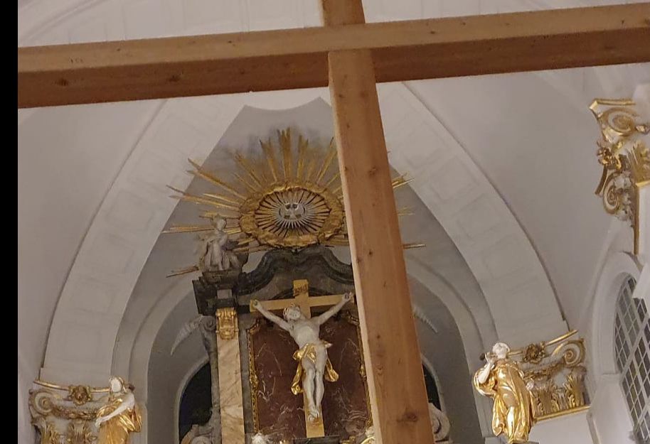 Innenansicht einer Kirche mit einem großen Holzkreuz im Vordergrund und einem kunstvoll verzierten Altar im Hintergrund.