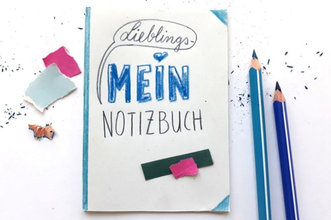 Ein Notizbuch mit der Aufschrift 'Lieblings-Mein Notizbuch' liegt neben zwei blauen Buntstiften und bunten Papierschnipseln.