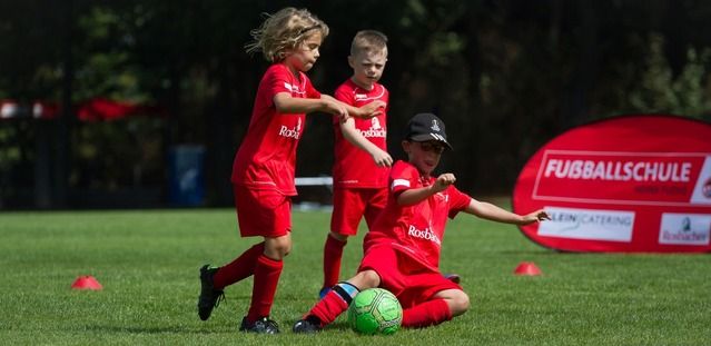 Drei Kinder in roten Fußballtrikots spielen auf einem Fußballfeld. Ein Kind sitzt am Boden mit einem Ball, während die anderen beiden stehen.