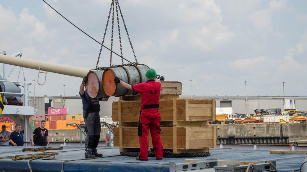 Arbeiter verladen Fässer und Kisten auf einem Schiff im Hafen.
