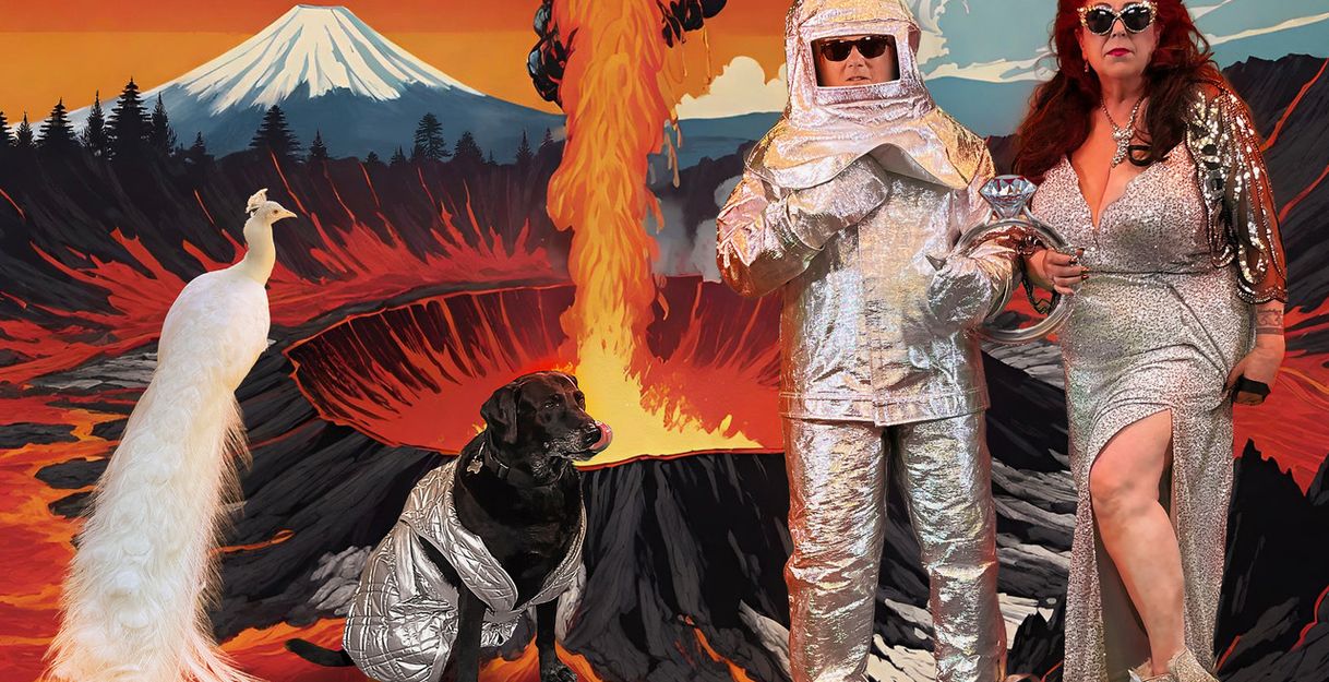 Two people in space outfits stand together with a dog and a white peacock in front of an illustrated background. This shows a natural landscape with a mountain, trees, and an erupting volcano.
