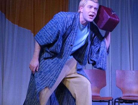 Ein Schauspieler in einem blauen Kimono und beigen Hosen hält einen roten Koffer ans Ohr auf einer Bühne.