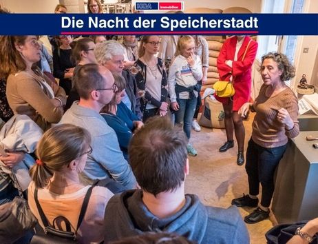 Eine Gruppe von Menschen hört einer Frau in einem Raum mit Kaffeesäcken zu. Oben steht 'Die Nacht der Speicherstadt'.
