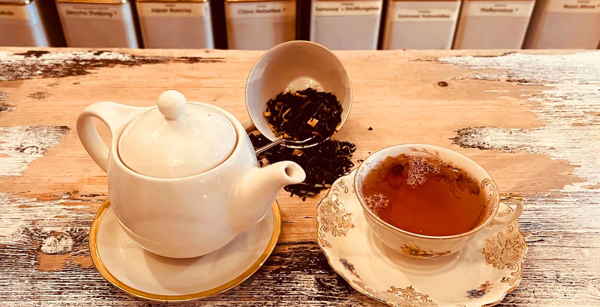 Eine Teekanne und eine Tasse Tee auf einem Holztisch, umgeben von Teeblättern und Teedosen im Hintergrund.
