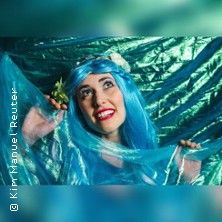 Frau mit blauer Perücke und Schleier, lächelnd vor einem schimmernden Hintergrund.