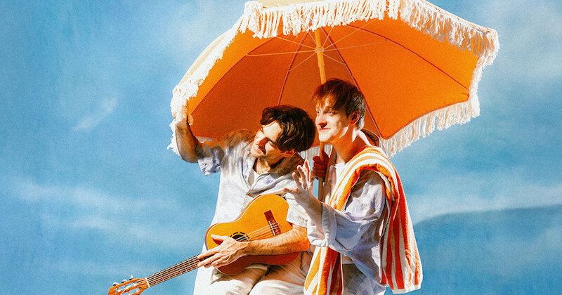 Zwei Männer unter einem orangefarbenen Sonnenschirm, einer hält eine Gitarre. Sie tragen helle Kleidung und stehen vor einem blauen Himmel.