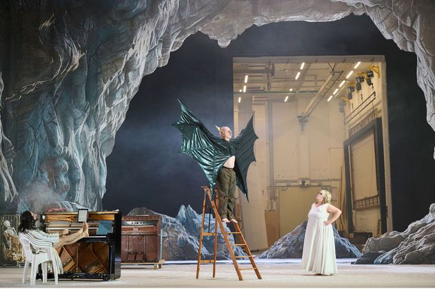 Bühnensituation. Ein Mann auf einer Leiter, die Arme zur Seite ausgestreckt, er trägt ein Kostüm mit großen schwarzen ledrigen Flügeln. Am Boden eine Frau im langen weißen Kleid, sie schaut zu ihm hoch. Links ein Mann am Klavier, die Füße auf die Tasten gelegt. Im Hintergrund eine Höhlenlandschaft. Die offene Hinterbühne ist zu sehen.