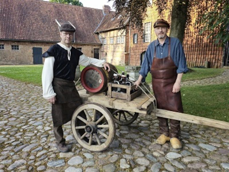 Zwei Männer in historischer Kleidung stehen neben einem Holzwagen mit einem Fass auf einem gepflasterten Hof.