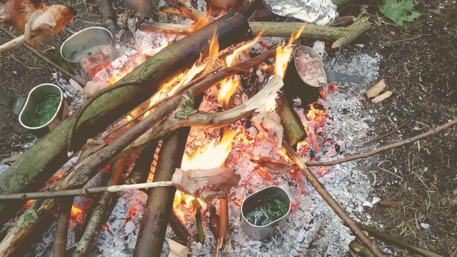 Lagerfeuer mit Kochutensilien und Fleisch am Spieß.