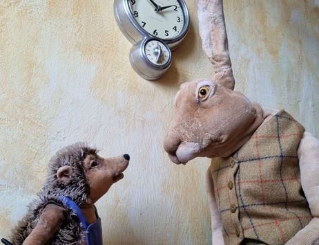 Ein Plüschhase und ein Plüschigel stehen vor einer Wanduhr und schauen sich an.