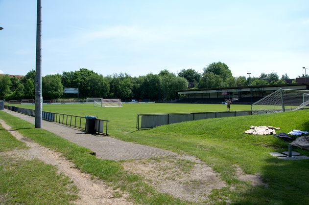 Fußballstadion mit leeren Tribünen und grünem Spielfeld, umgeben von Bäumen.
