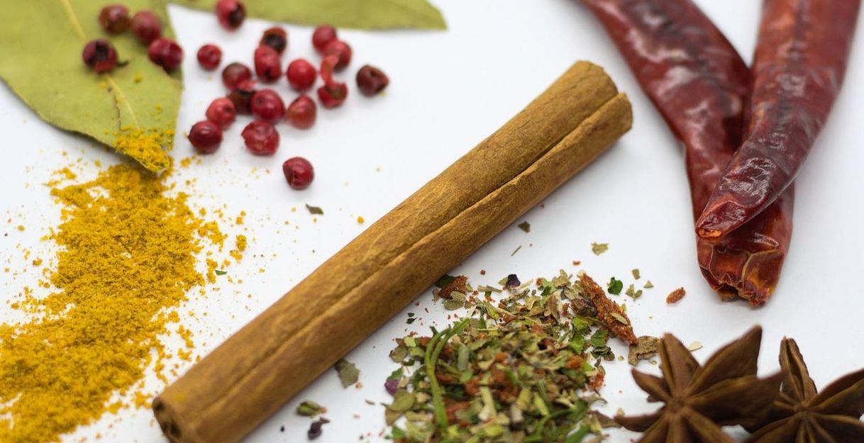 Verschiedene Gewürze auf weißem Hintergrund, darunter Zimtstange, Lorbeerblätter, rote Beeren, getrocknete Chilischoten und Sternanis.