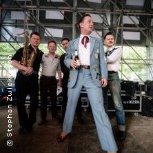 Eine Band posiert auf einer Bühne mit Musikinstrumenten im Hintergrund.