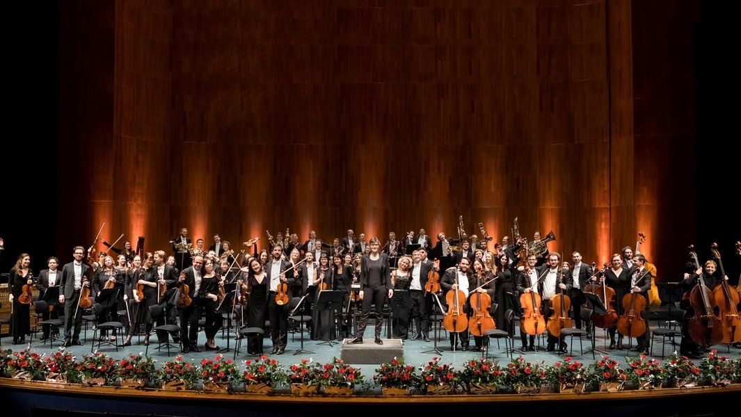 Ein Orchester auf einer Bühne mit Blumen geschmückt, Musiker stehen mit Instrumenten und verbeugen sich vor dem Publikum.
