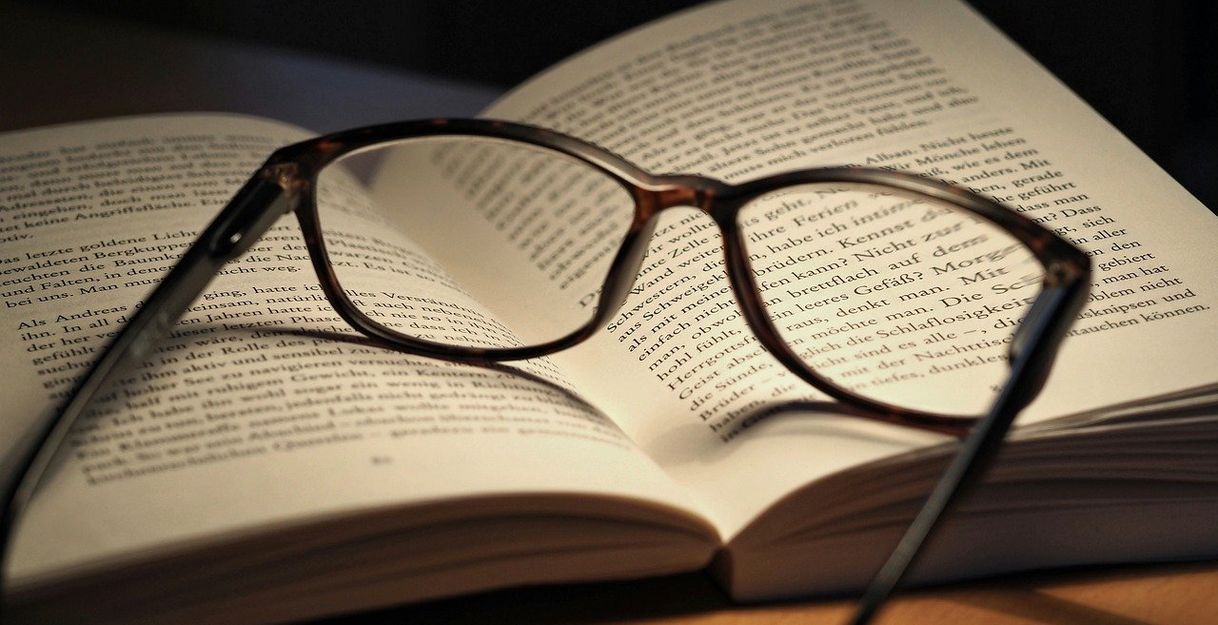 Eine Brille liegt auf einem aufgeschlagenen Buch, das auf einem Tisch liegt.