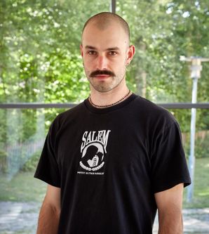 Einjunger Mann mit Schnauzer und Halbglatze in einem schwarzen Tshirt schaut direkt in die Kamera