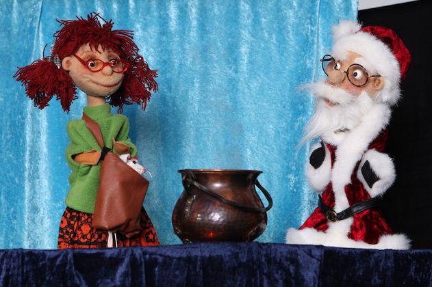 Zwei Puppen, eine als Weihnachtsmann und eine mit roten Haaren, stehen vor einem Kupferkessel auf einer Bühne.