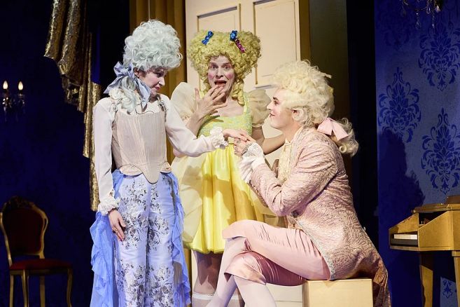 Drei Schauspieler in barocken Kostümen auf einer Bühne, einer kniet vor den anderen.