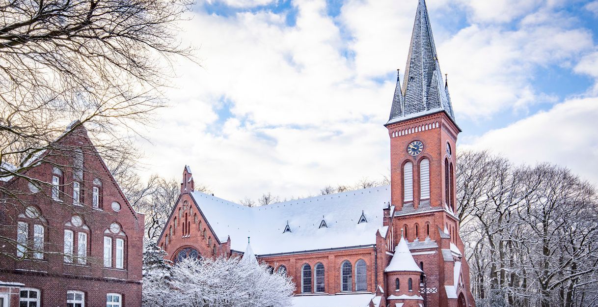 Eine rote Backsteinkirche im Winter mit schneebedecktem Dach und Turm, umgeben von schneebedeckten Bäumen und einem blauen Himmel.