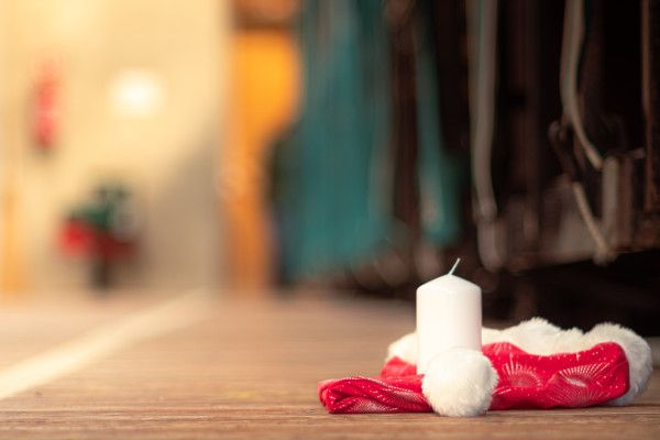 Weiße Kerze mit rotem Nikolausmütze-ähnlichem Stoff und weißem Bommel auf einem Holzboden; im unscharfen Hintergrund ein Innenraum.