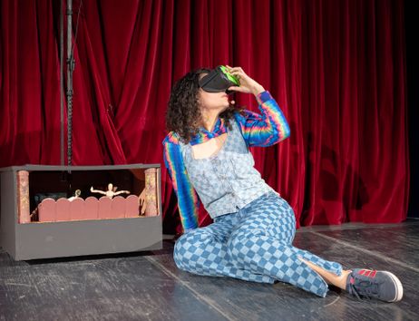 A woman dressed in colourful clothes is sitting on a stage in front of a red velvet curtain, holding a pair of VR glasses up to her eyes.