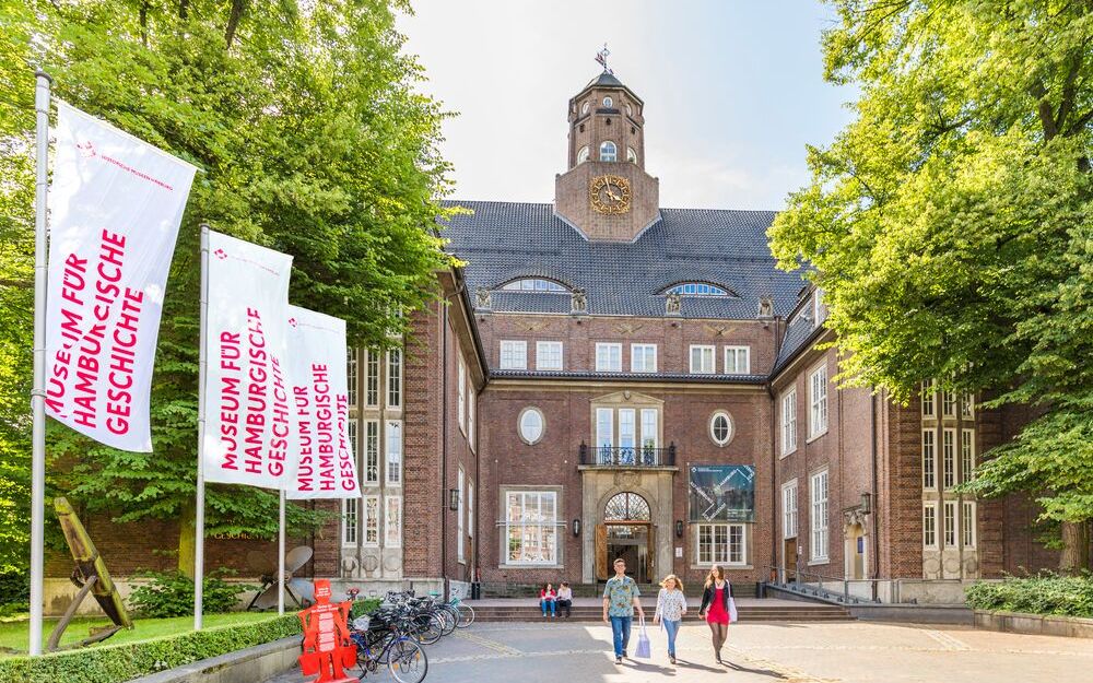 Menschen vor einem palastähnlichen Gebäude. Der rote Backsteinbau geht über 5 Etagen. Der Eingangsbereich ist über Treppen erreichbar, hat ein steinernes Tor und ist von Fenstern umrandet. Darüber ein Balkon. Auf dem Dach ist mittig ein Turm.